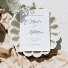 A white save the date card on both a marble background and a white seashell style plate alongside neutral fabric
