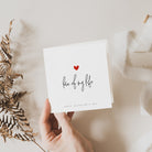 A person holding a Valentine's Day card with the phrase 'love of my life' written on it, accompanied by a small red heart illustration. The card is presented against a neutral background with some dried plants to the side.