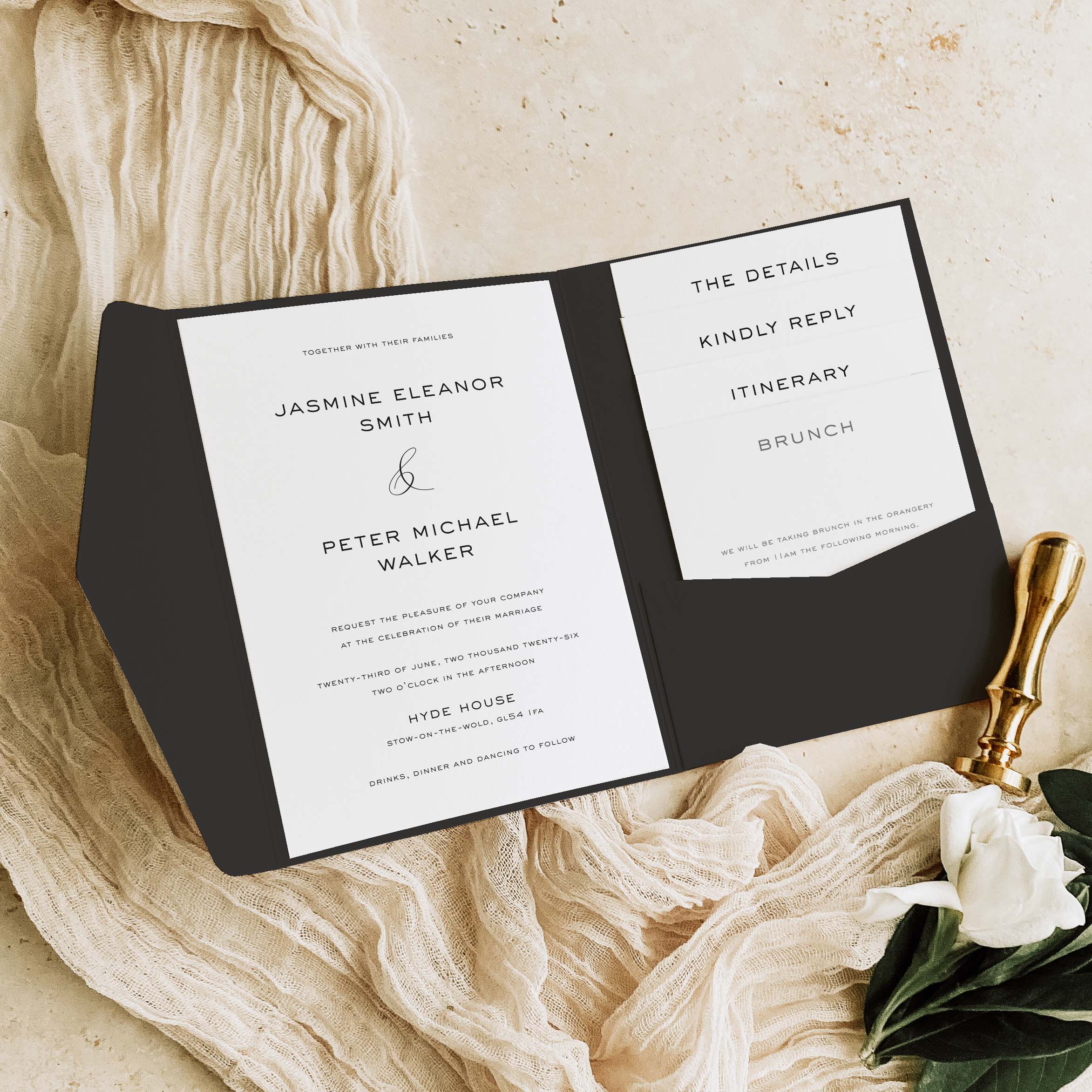 Pocketfold Wedding invitation set on a textured surface with a gold pen and white flowers.