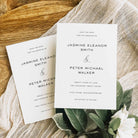 Two white save the date cards for a wedding with elegant text, shown on a wooden board with white fabric and green foliage.