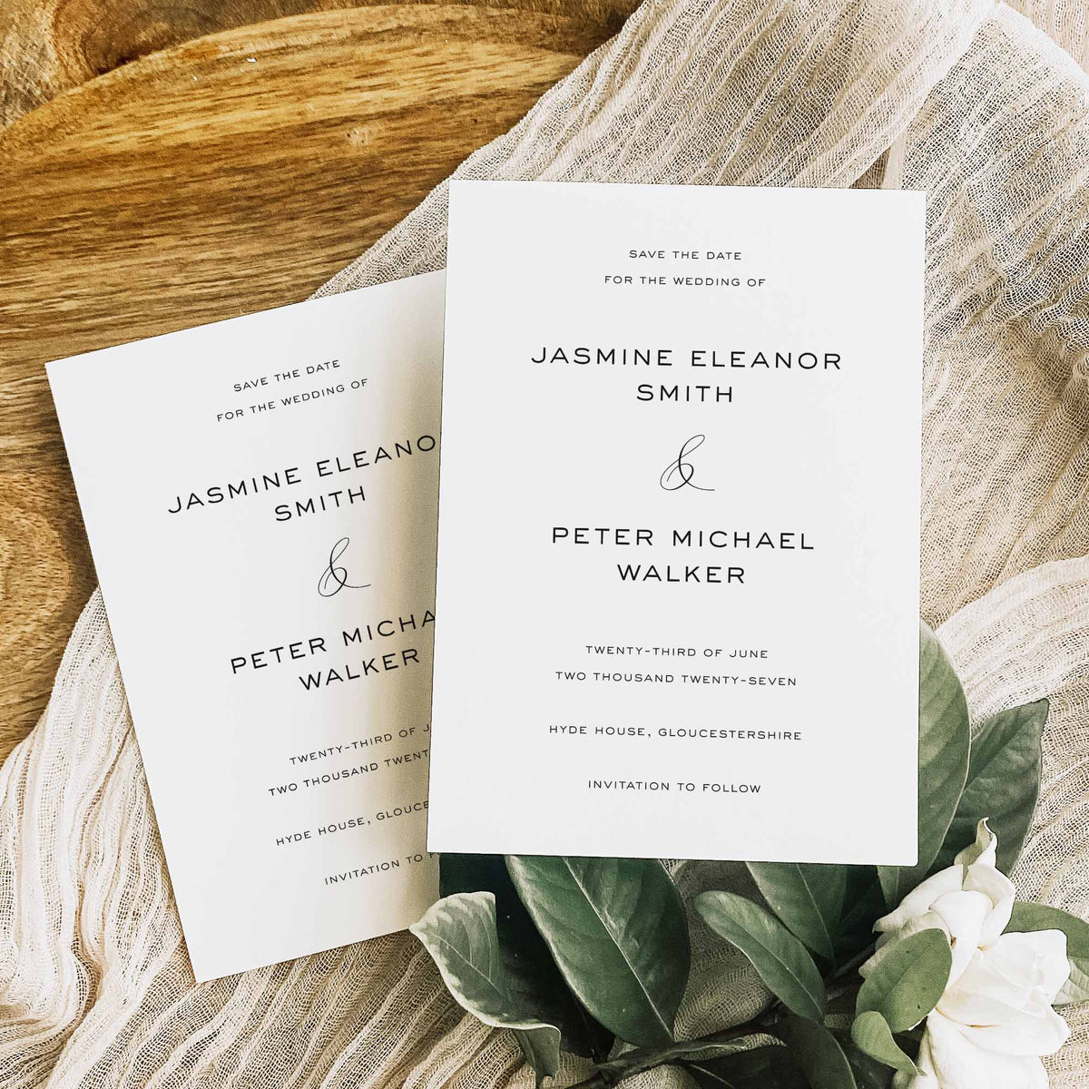 Two white save the date cards for a wedding with elegant text, shown on a wooden board with white fabric and green foliage.