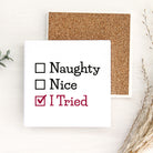 A white square sign or magnet with printed text reading 'Naughty', 'Nice', and 'I Tried' with checkboxes, styled on a light wood surface with a cork board behind it and dried grasses at the side.
