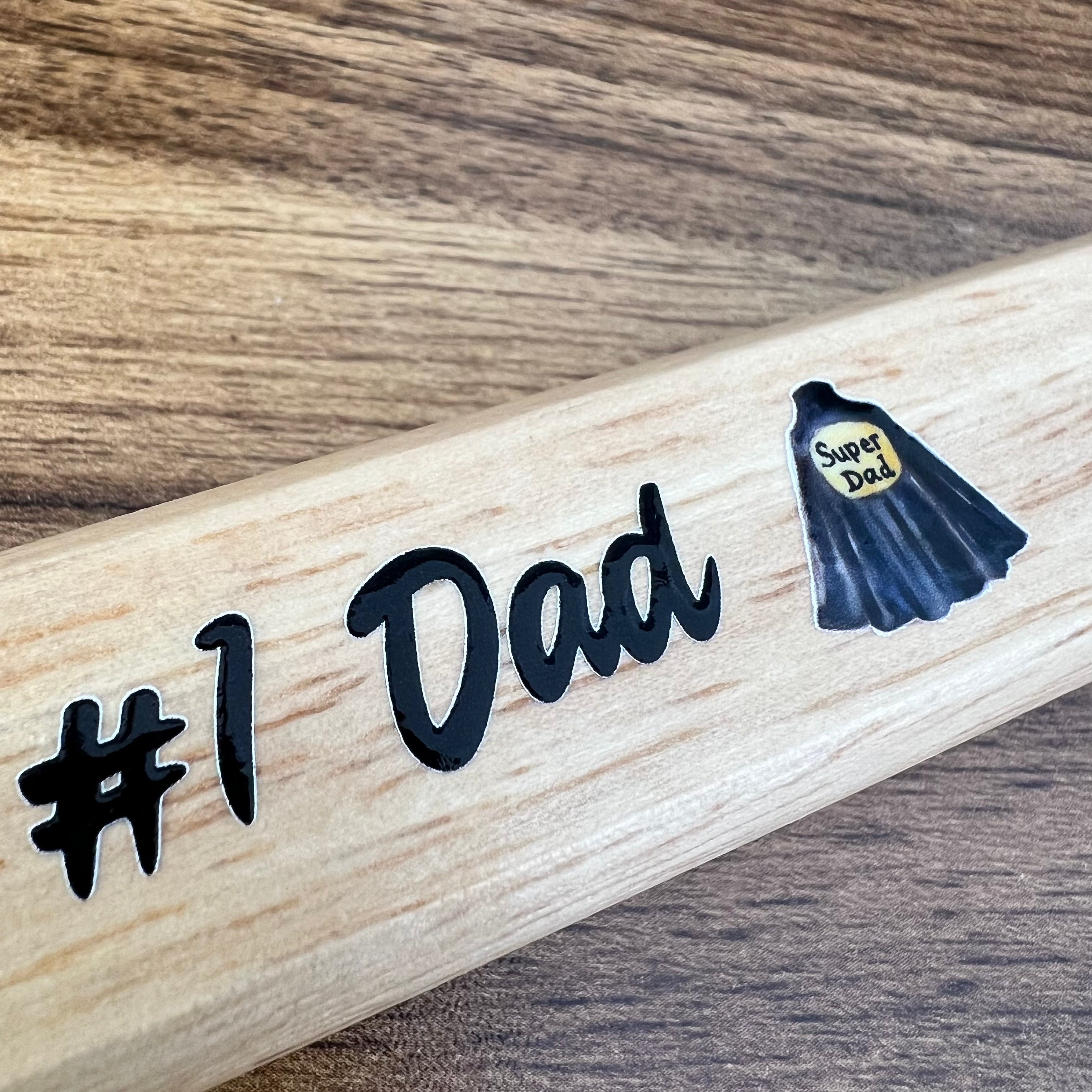 Wooden bat with '#1 Dad' and 'Super Dad' stickers on a wooden surface