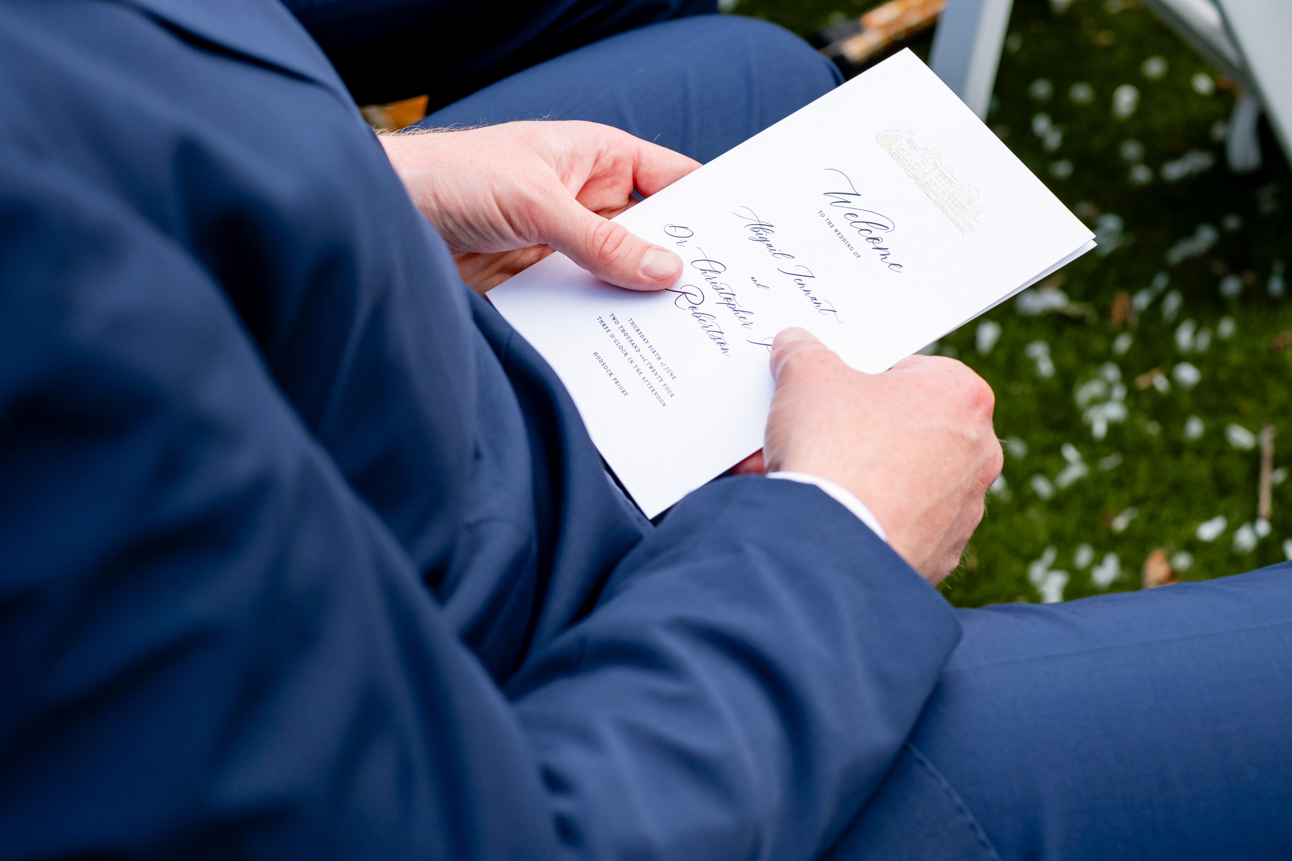 Person holding a wedding program with gold foil venue sketch and grass background