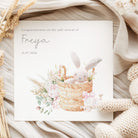 NEW BABY  card with a bunny in a basket and floral elements on a soft fabric background.