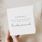 Hand holding a card with 'Harriet, get your glam face on, it's time to be my bridesmaid' text.