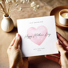 Person holding a Mother's Day card with a pink heart design on a wooden surface.