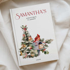 A personalized 2026 Christmas planner titled 'Samantha's Christmas Planner' resting on a soft, draped cream fabric. The cover is white with elegant red script text and a watercolor illustration of a decorated Christmas tree, a red cardinal, a mailbox, and holly leaves.