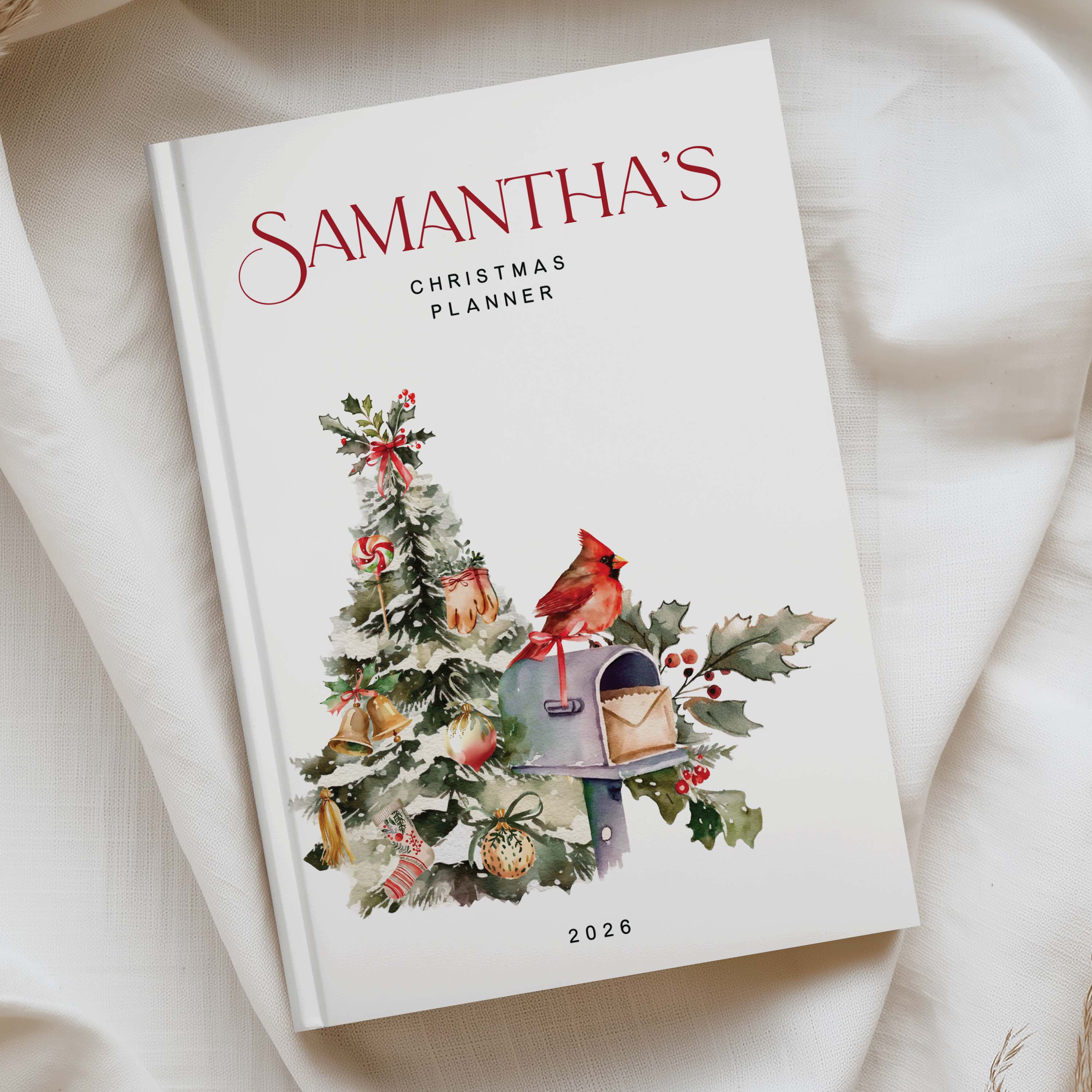 A personalized 2026 Christmas planner titled 'Samantha's Christmas Planner' resting on a soft, draped cream fabric. The cover is white with elegant red script text and a watercolor illustration of a decorated Christmas tree, a red cardinal, a mailbox, and holly leaves.