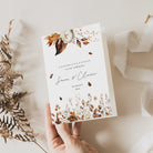 A hand holding a floral wedding day card with autumnal leaves design, placed next to some dried plants on a white textured surface.