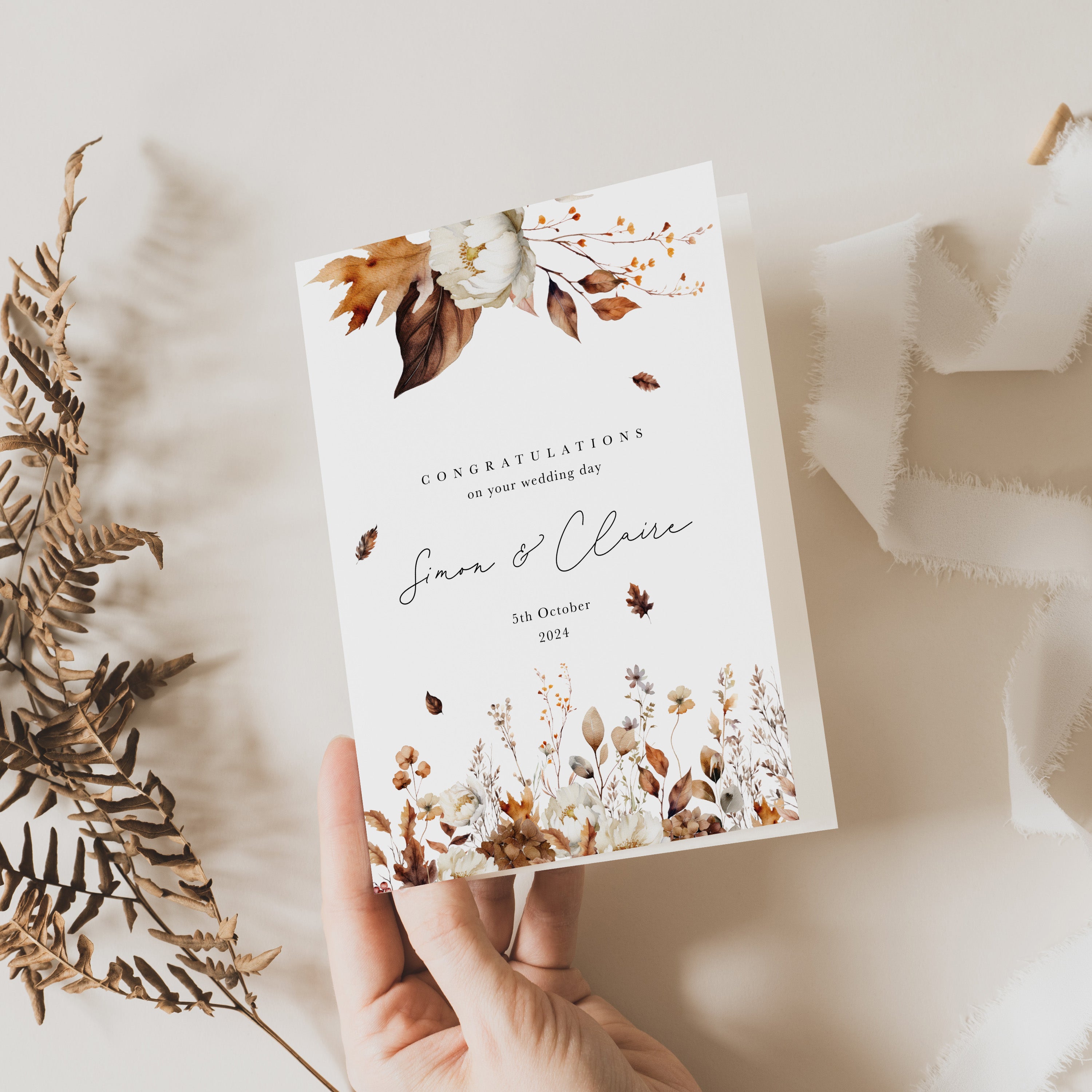 A hand holding a floral wedding day card with autumnal leaves design, placed next to some dried plants on a white textured surface.
