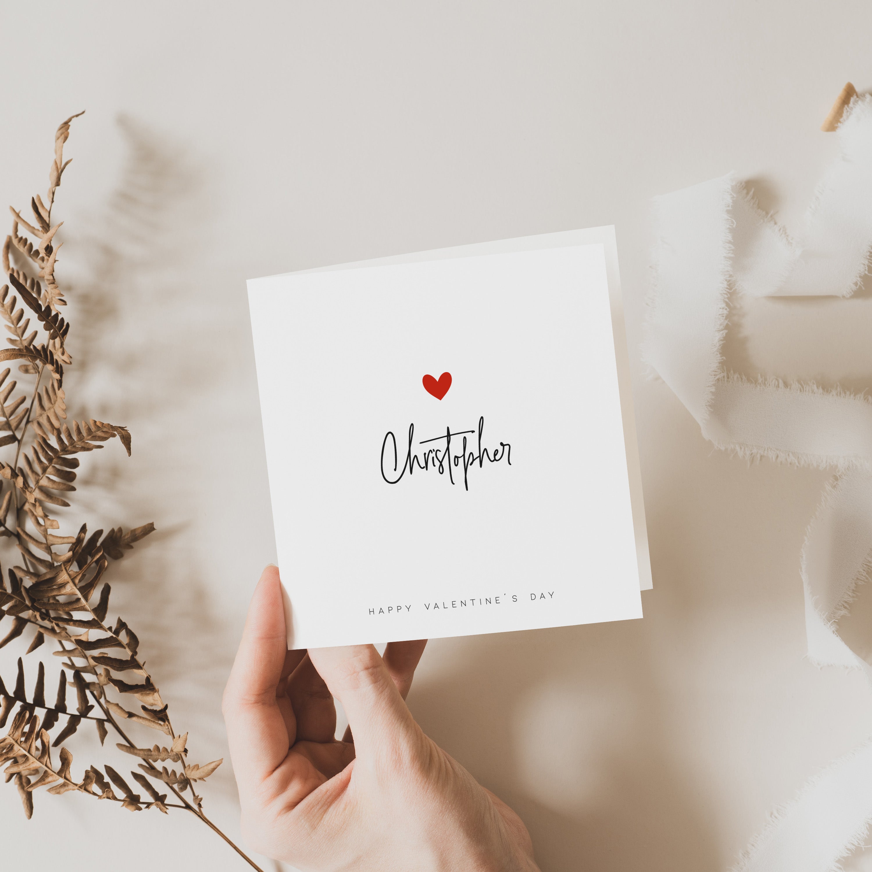 A personalized Valentine's Day card with the name 'Christopher' printed on it, held in a hand, decorated with a small red heart, presented on a neutral background with some foliage to the side.