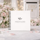 White spiral-bound guestbook with personalised text on a decorative background with flowers and cards.