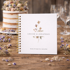 Wedding guest book with floral design and text on a wooden table with flowers and glasses.