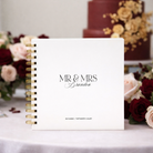 White guestbook with 'MR & MRS Brandon' on a table with flowers and a cake in the background