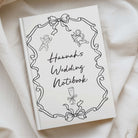 A hardcover wedding notebook titled 'Hannah's Wedding Notebook' in black script, decorated with line-art illustrations of cherubs, ribbons, and a champagne glass. The notebook is photographed on draped cream fabric.