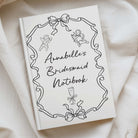 A white hardcover notebook with black line art illustrations and the title 'Annabelle's Bridesmaid Notebook' in script, photographed on draped cream fabric.