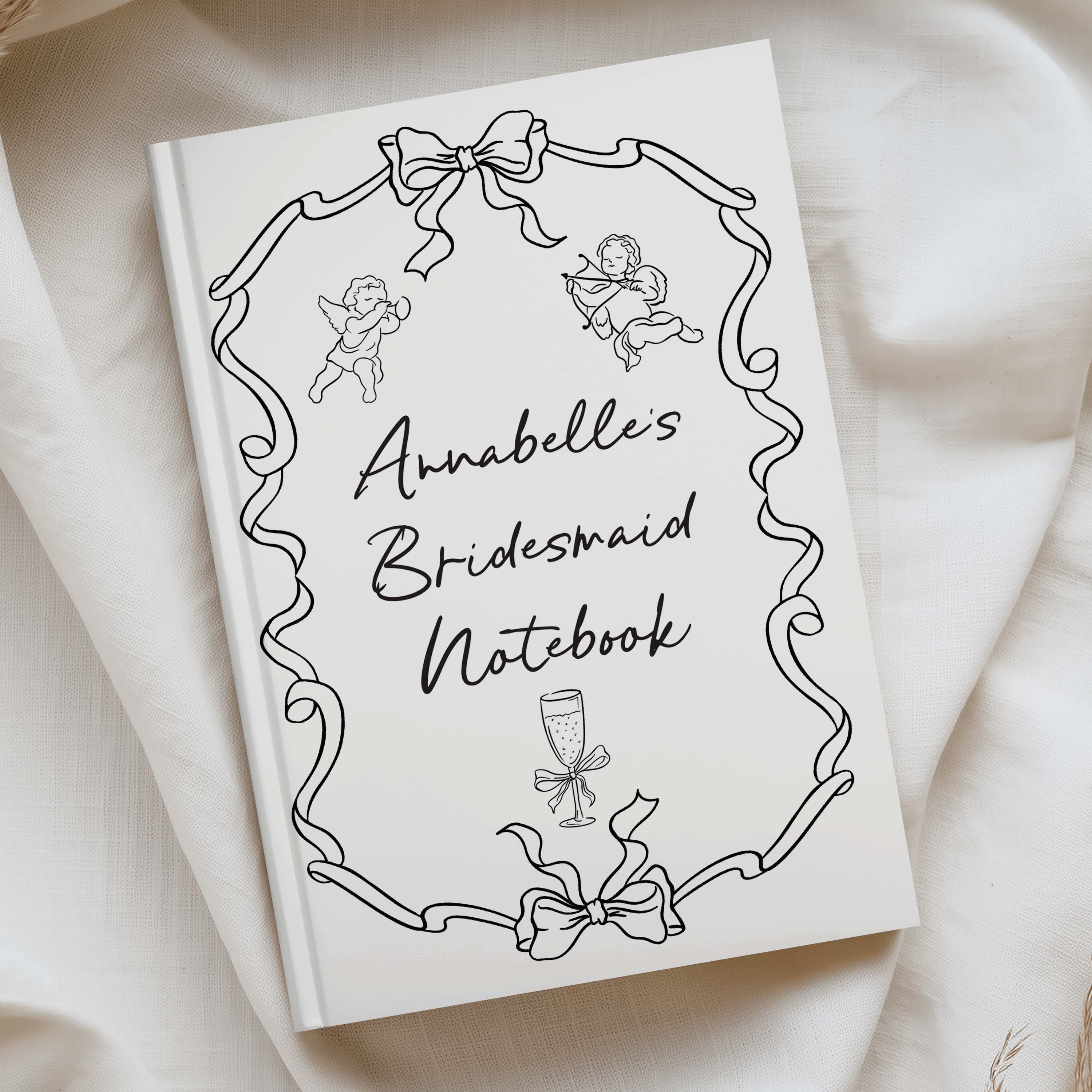 A white hardcover notebook with black line art illustrations and the title 'Annabelle's Bridesmaid Notebook' in script, photographed on draped cream fabric.