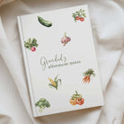 A hardcover notebook titled 'Grandad's allotment notes' with watercolor illustrations of various vegetables, styled on a draped cream-colored fabric background.