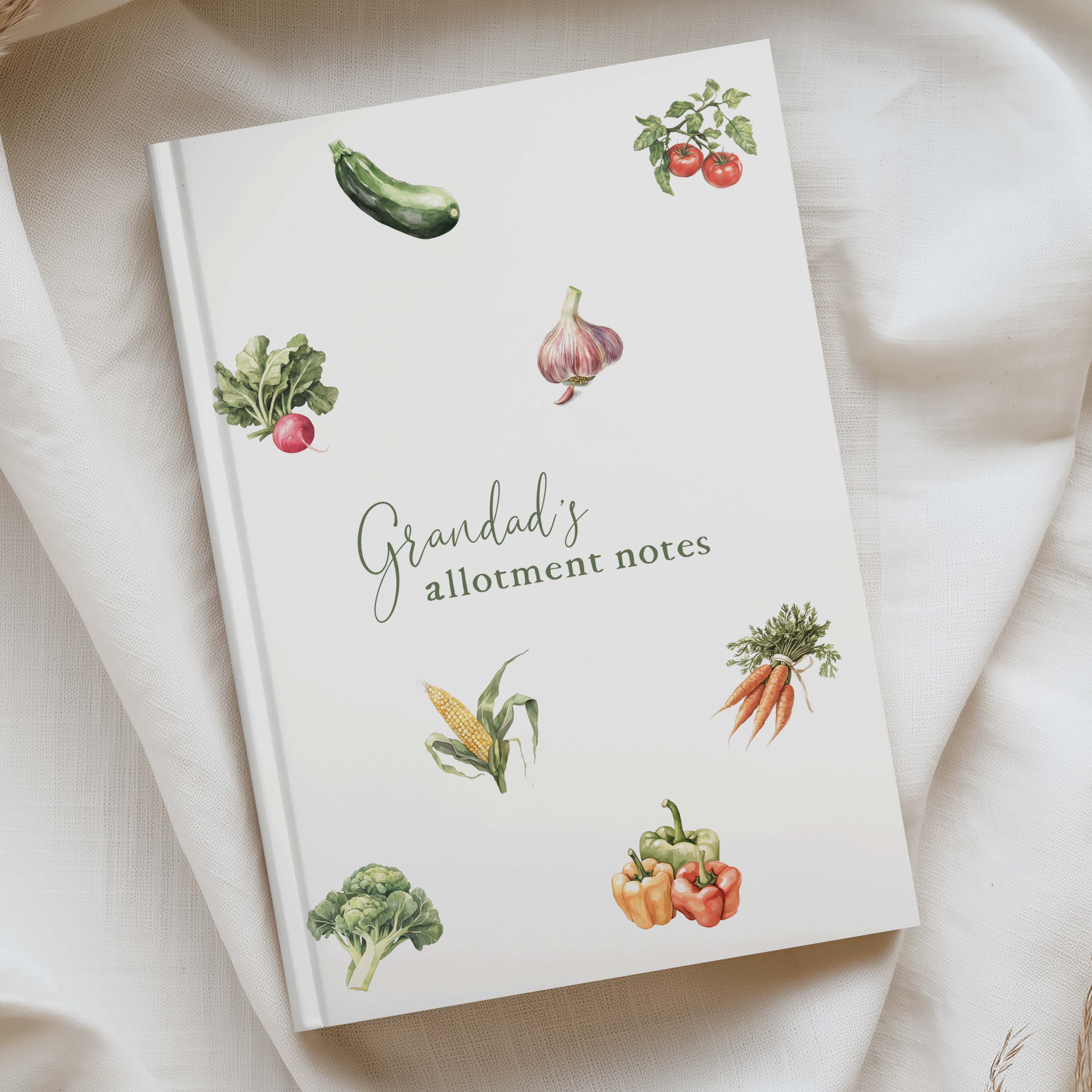 A hardcover notebook titled 'Grandad's allotment notes' with watercolor illustrations of various vegetables, styled on a draped cream-colored fabric background.
