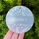 A round frosted glass Christmas ornament with white botanical illustrations and the name 'HARRIET' in the center, held by a hand against a background of green pine branches.