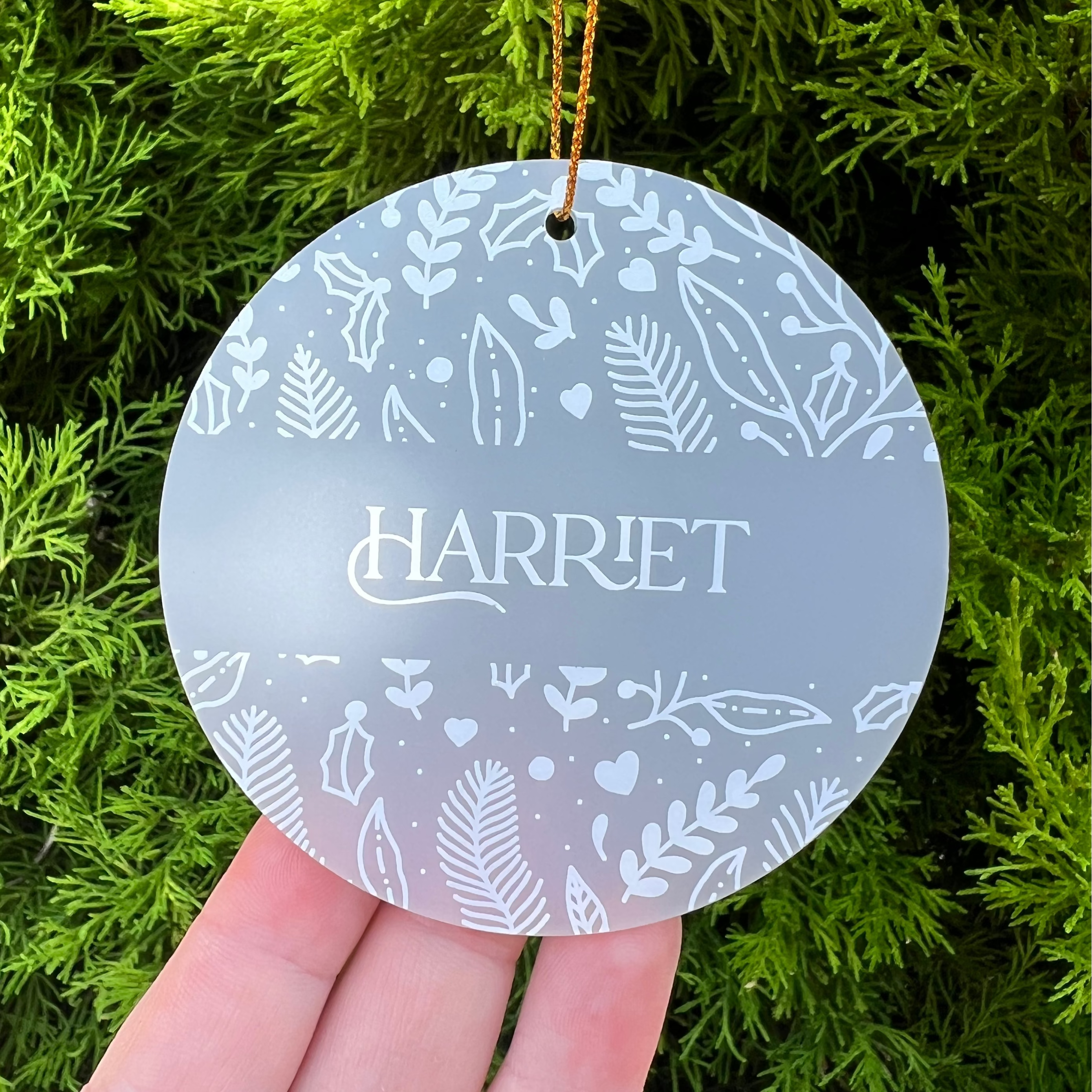 A round frosted glass Christmas ornament with white botanical illustrations and the name 'HARRIET' in the center, held by a hand against a background of green pine branches.