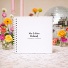 White guestbook with 'Mr & Mrs Roland' text, surrounded by colorful flowers and a disco ball.