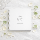 white wedding guest book with white binder, laid flat on a white tablecloth with green and white petals