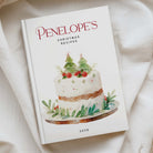 A hardcover cookbook titled 'PENELOPE'S CHRISTMAS RECIPES 2026' resting on a draped white fabric background. The cover features a watercolor illustration of a decorated Christmas cake with green holly leaves and red berries.
