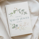 A personalized wedding guest book or notebook titled 'MAIDLINE'S MAID OF HONOUR NOTEBOOK' with delicate watercolor floral illustrations, displayed on a soft beige fabric background.