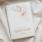 A personalized bridesmaid notebook with a watercolor floral design in pink and green on a white cover, styled on a draped cream fabric background.