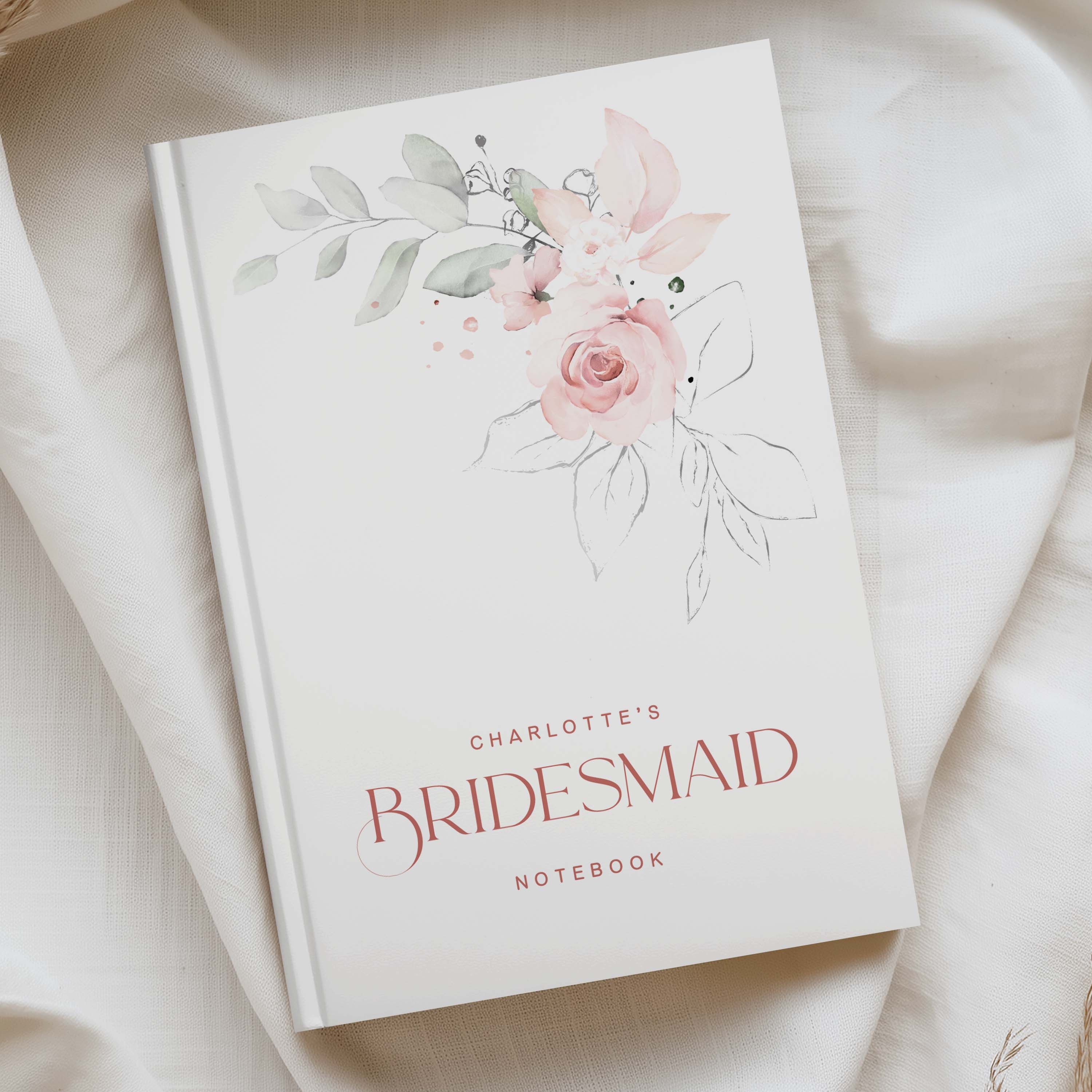 A personalized bridesmaid notebook with a watercolor floral design in pink and green on a white cover, styled on a draped cream fabric background.