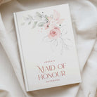 White a5 notebook with pink and green watercolour florals and the phrase "FREYAS MAID OF HONOUR NOTEBOOK" IN RICH PINK TEXT