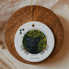 A round white memorial ornament with a hanging loop, featuring a custom photo of a black dog surrounded by greenery and floral accents, with the text 'YOU LEFT PAW PRINTS ON OUR HEART' and the name 'BRUNO' below. The ornament is displayed on a round wooden tray against a soft beige fabric background.