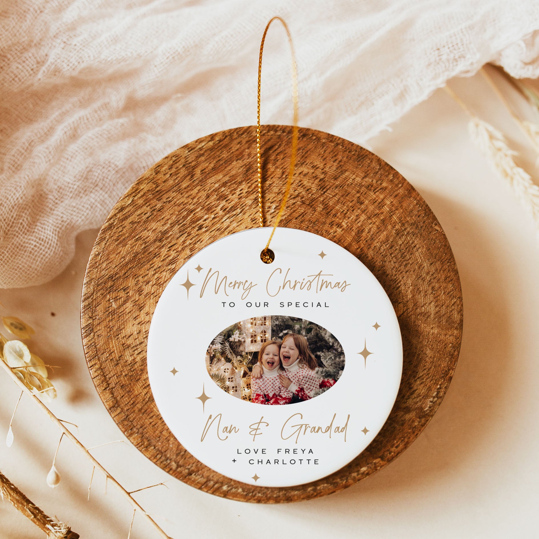 A personalized Christmas ornament displayed on a round wooden stand. The ornament is white with gold script text reading 'Merry Christmas to our special Non & Grandad' and a photo of a grandfather and grandmother, with the names 'Love Freya + Charlotte' below.