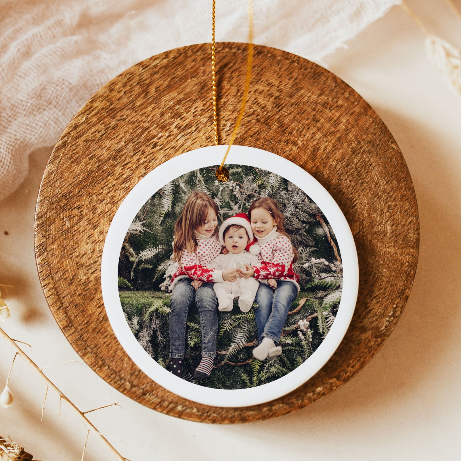 A round wooden Christmas ornament with a white inner border and a printed photo of three children sitting on a Christmas tree. The ornament hangs from a gold string and is displayed on a neutral beige surface with soft fabric and dried floral accents.