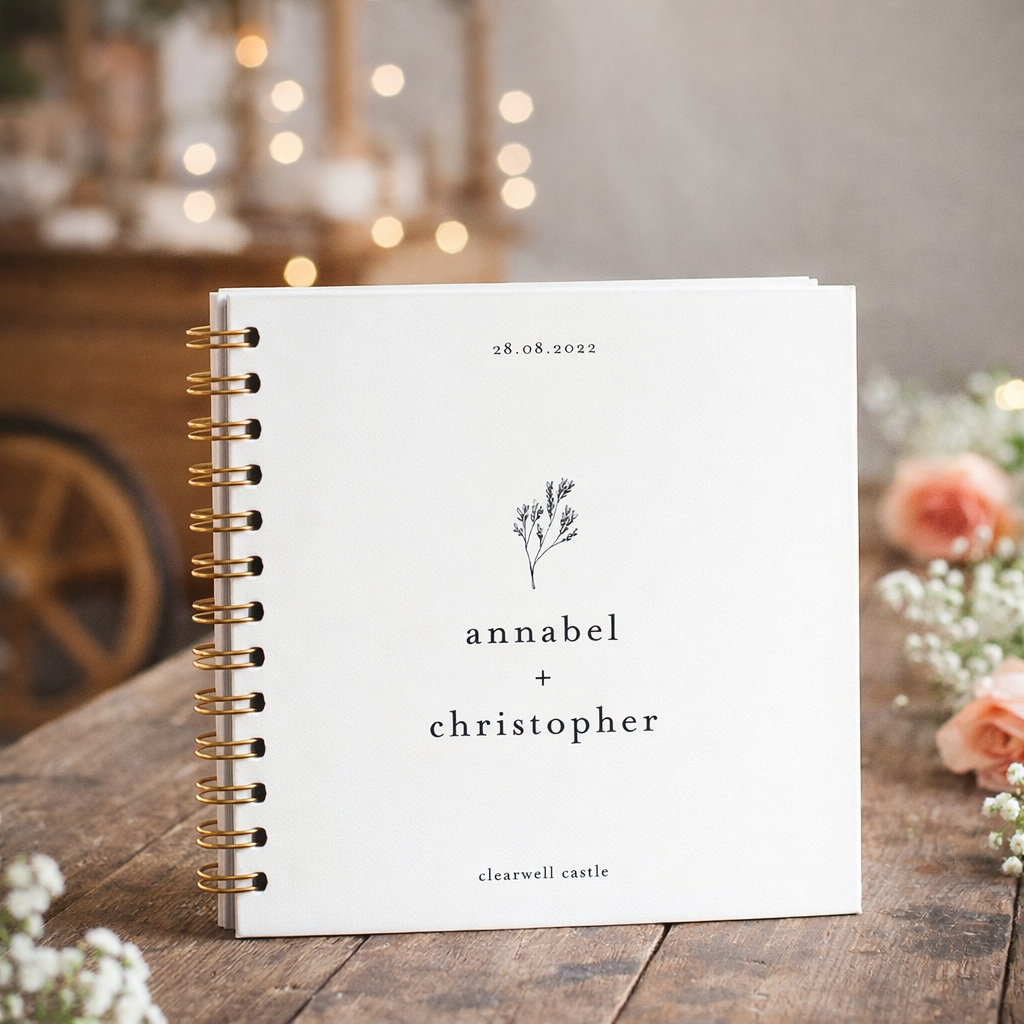 White guestbook with gold spiral binding on a wooden surface with blurred background