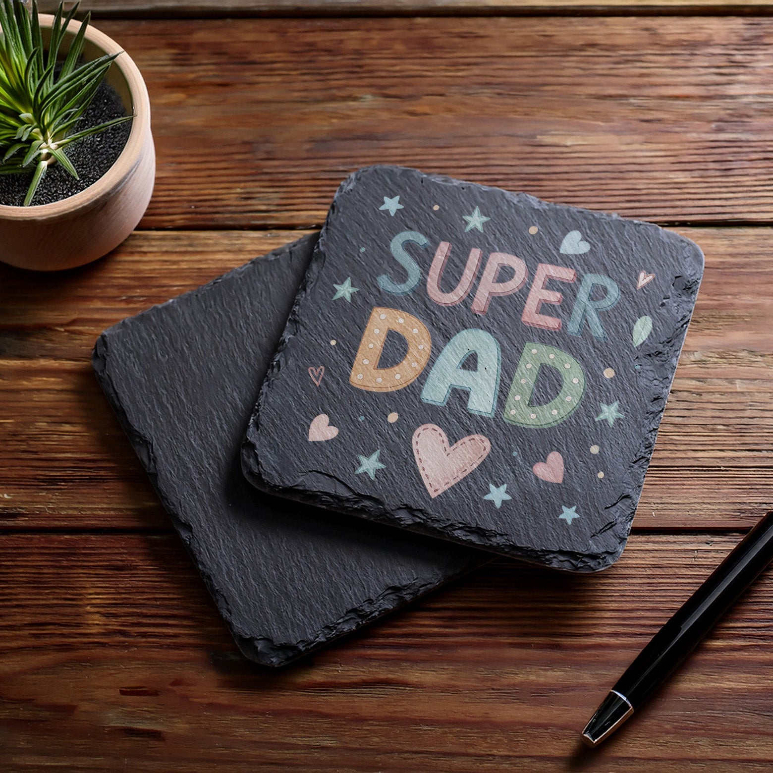 Slate coaster with 'Super Dad' text on a wooden surface with a pen and plant.