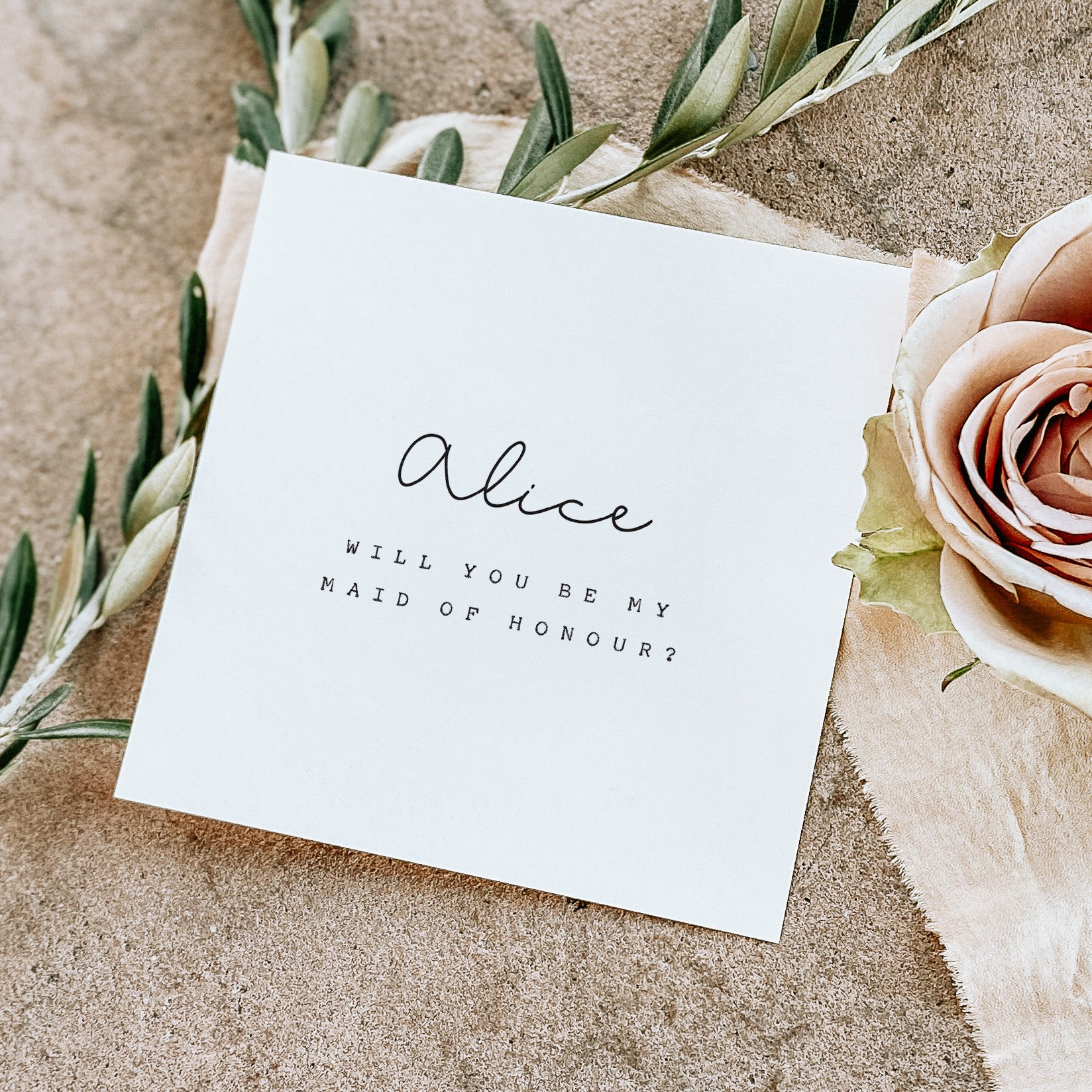 White card with 'Will you be my Maid of Honour?' text on a textured surface with flowers.