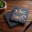 Slate coaster with 'King of Dads' text on a wooden surface with a plant and pen.