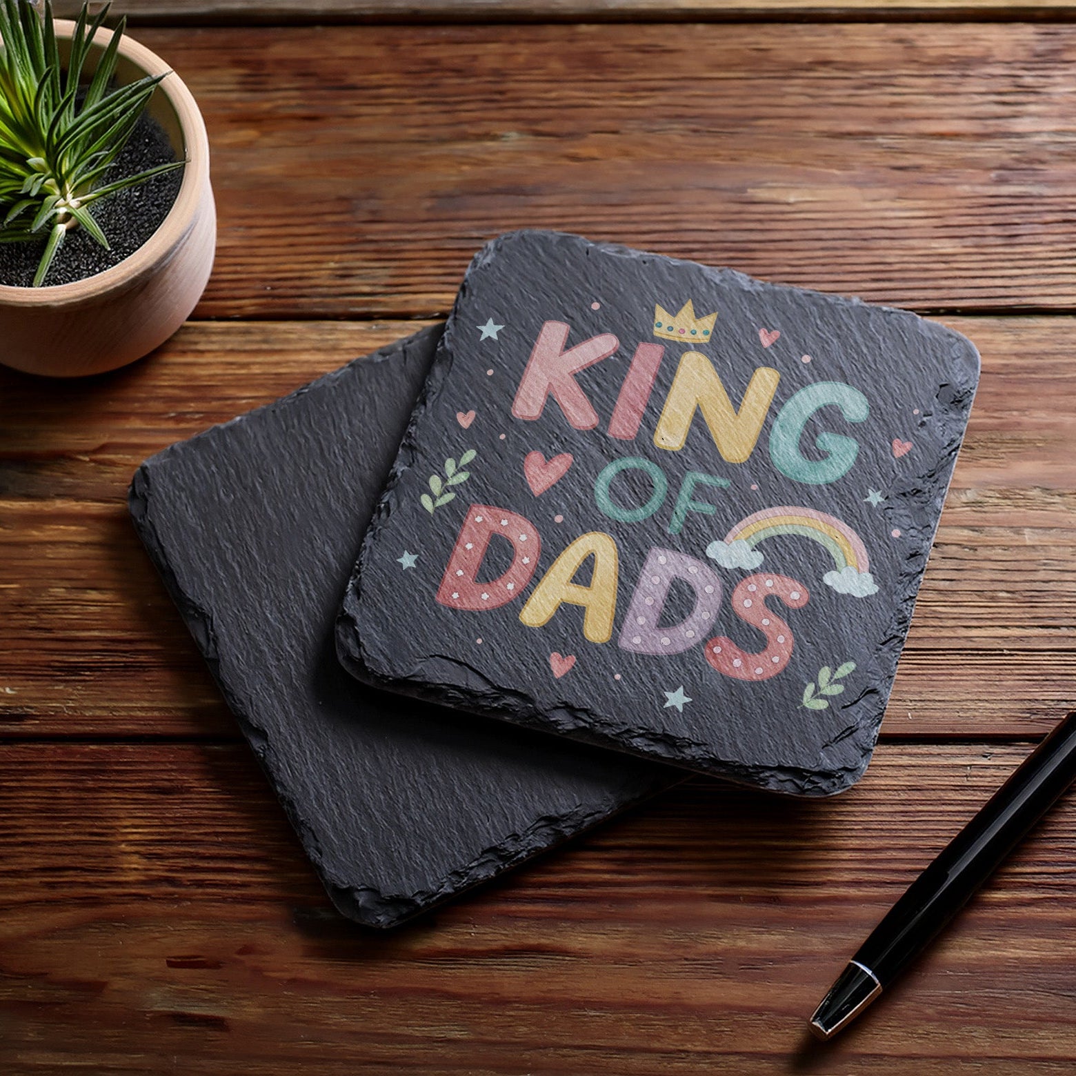 Slate coaster with 'King of Dads' text on a wooden surface with a plant and pen.