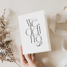 A hand holding a modern and abstract wedding day card with the word 'wedding' written in bold black letters, presented on a neutral background with some foliage decoration.