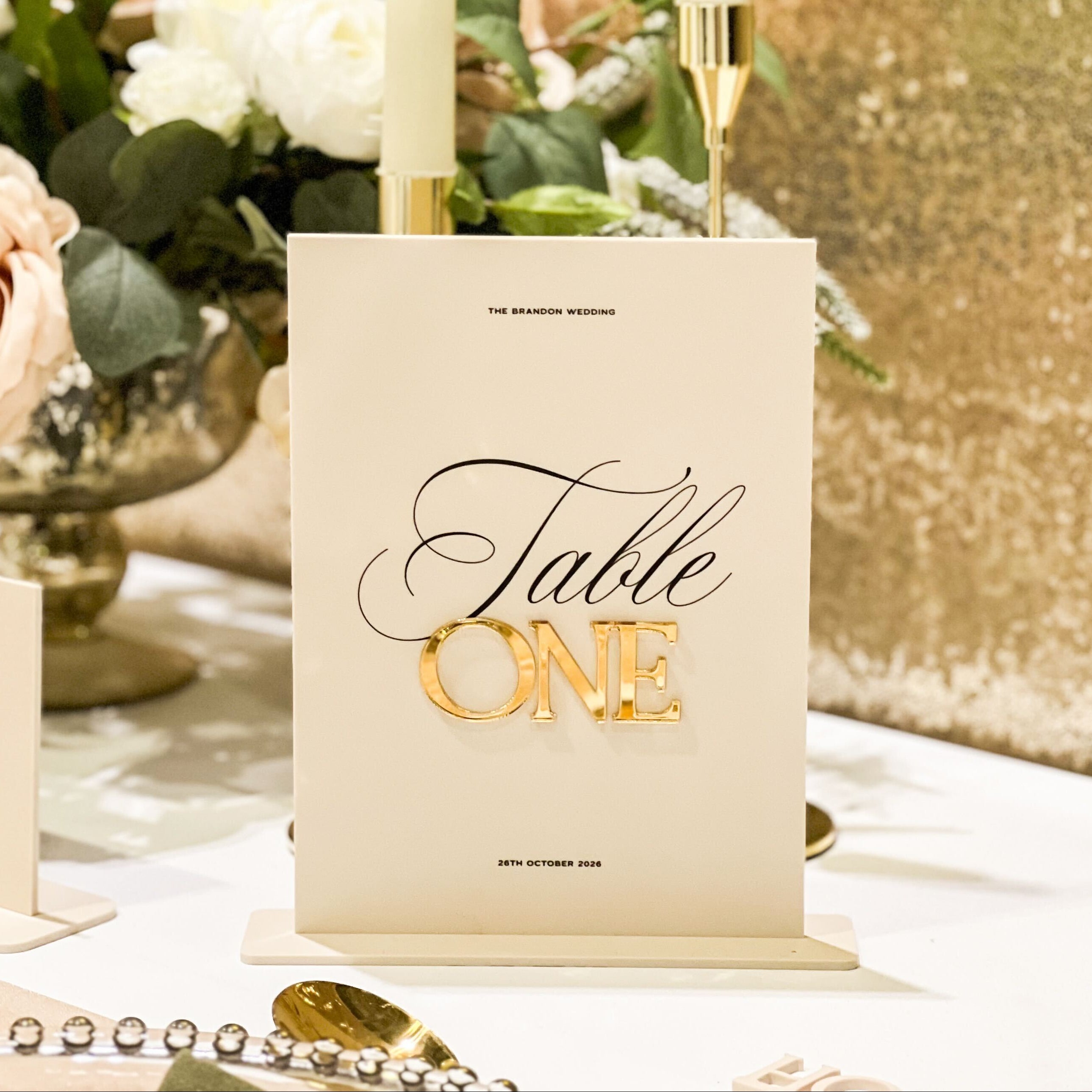 Table setting with 'Table ONE' sign, candles, and flowers on a gold sequin background