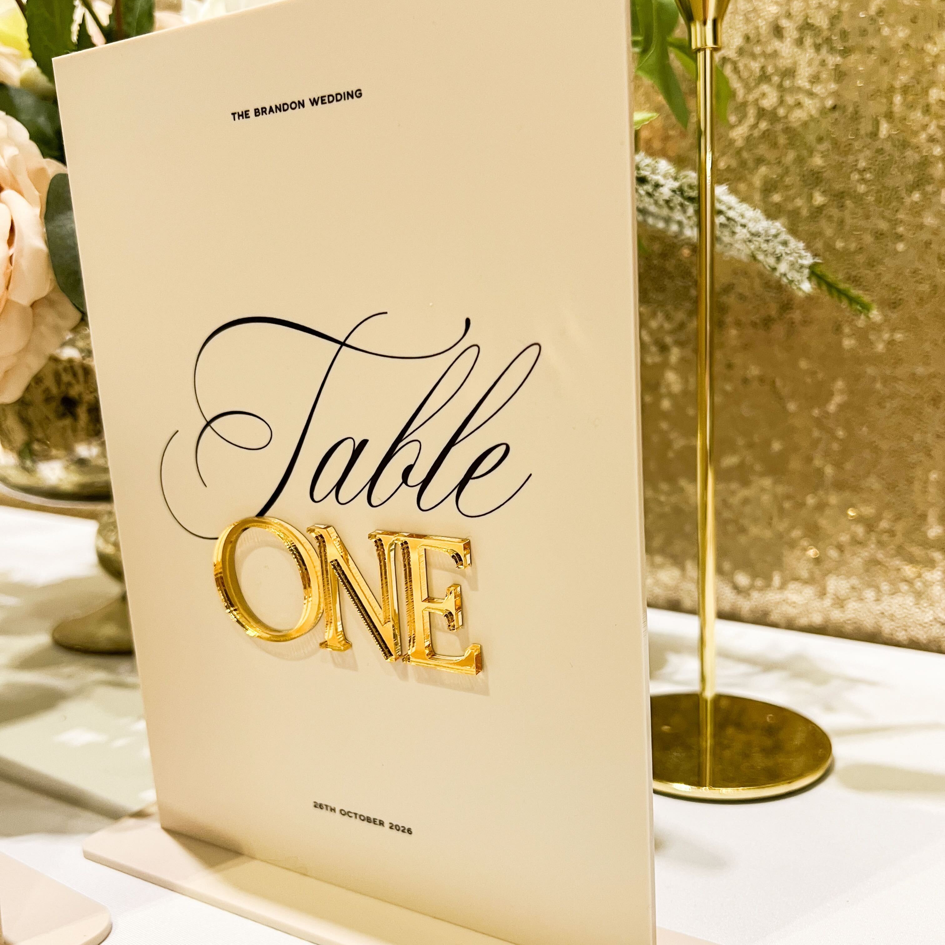 Table number card with 'Table One' in gold and black on a stand, with flowers in the background.