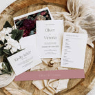 A styled flat-lay of wedding stationery on a rustic wood slice background with cream fabric and flowers. The set includes a floral invitation card, a white wedding invitation with 'Oliver & Victoria', an 'Order of the Day' card, and a 'Kindly RSVP' card on a dusty pink envelope.