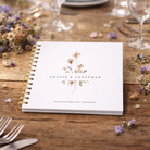 White guestbook with floral design and text on a wooden table with flowers and glasses.