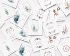 A flat-lay arrangement of multiple white Christmas greeting cards featuring delicate watercolor illustrations of Christmas trees, floral wreaths, pinecones, and holiday motifs on a light background.