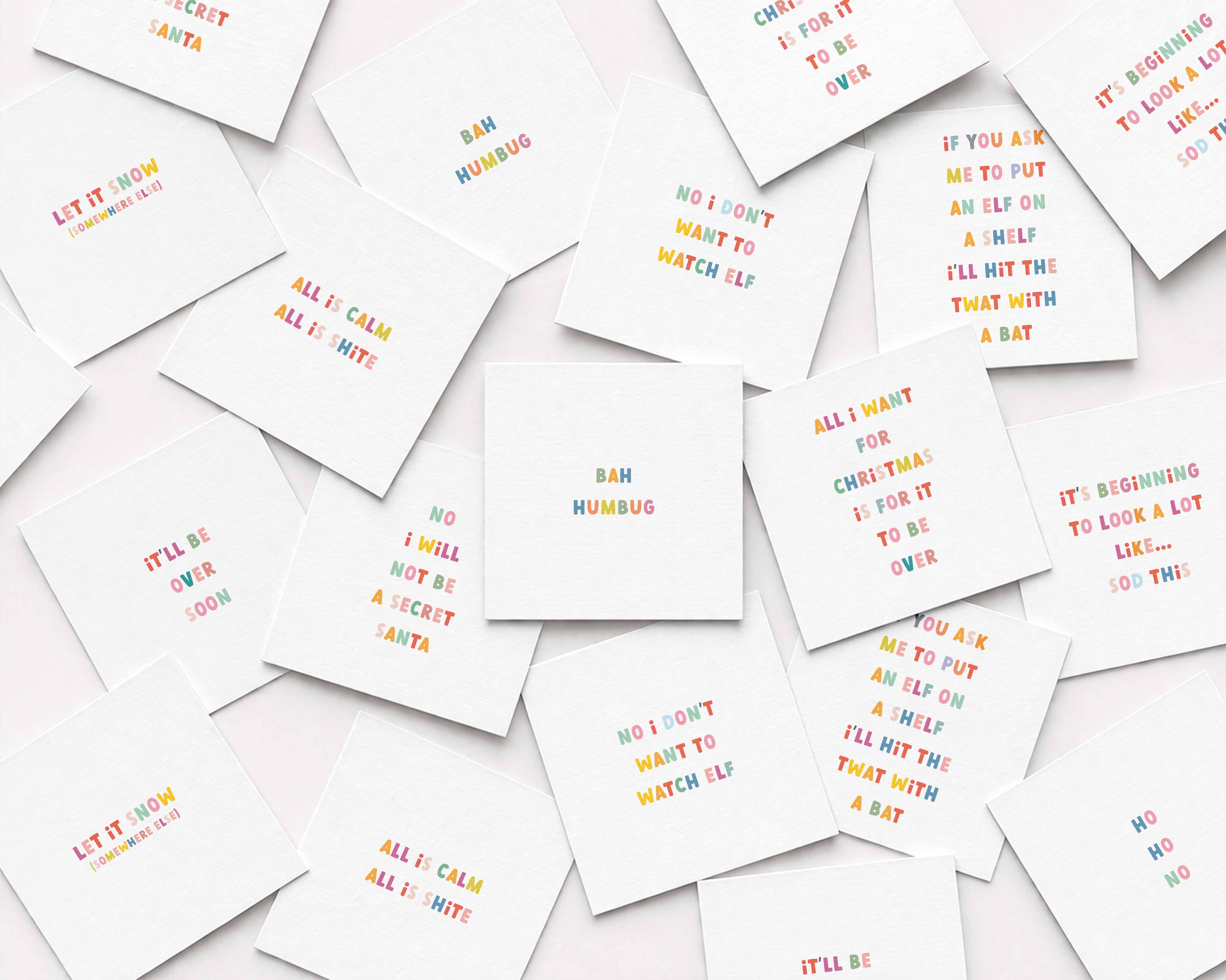 A scattered arrangement of small white square anti-Christmas cards printed with colorful playful text on a white background.