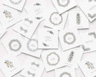 A scattered arrangement of white Christmas cards featuring delicate botanical illustrations in muted gray, blue, and yellow, including wreaths, sprigs, and small text such as Merry Christmas and Happy New Year.
