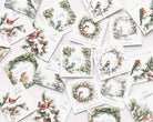 A flat-lay arrangement of multiple white Christmas greeting cards featuring watercolor illustrations of birds, wreaths, pine branches, and holiday text, photographed on a pale pink background.
